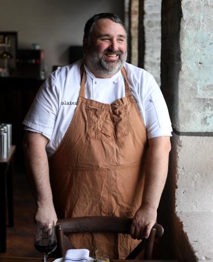 Photo of Chef Farrell Harrison in a brown apron, standing by a table looking out a window 