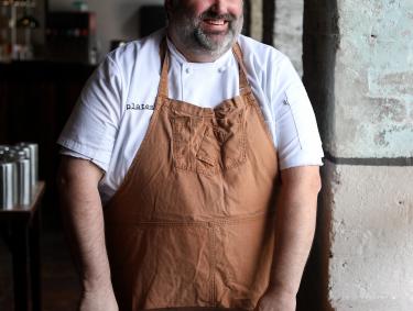 Photo of Chef Farrell Harrison in a brown apron, standing by a table looking out a window 